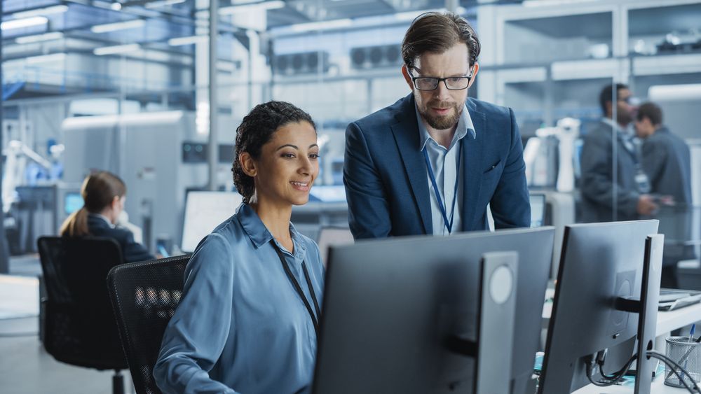 Portrait of a Young Male Team Leader and Hispanic Female Engineer Collaborating on a Project, Using Computer, Talking in a Factory with Robotic Arms Producing Modern Devices for Tech Industries.