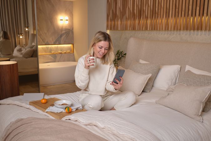 Happy female drinking coffee and browsing smartphone on bed