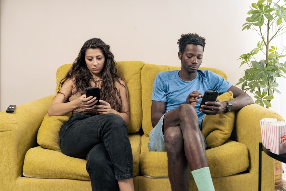 A young couple sits far apart on a couch, each staring silently at their own mobile phone, illustrating digital isolation.