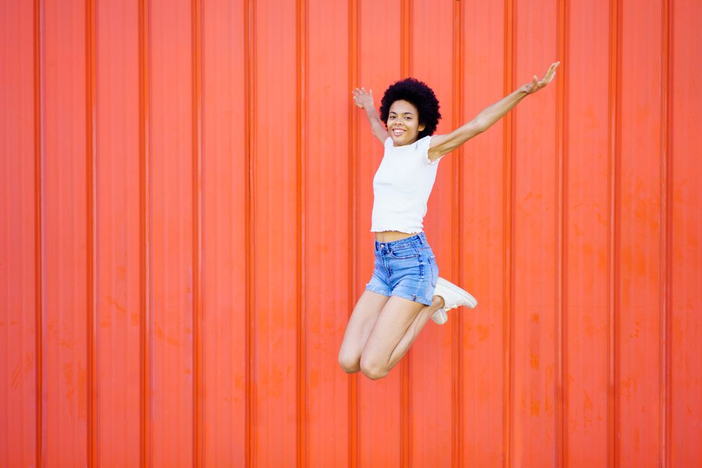 Happy woman in shorts and t-shirt jumping with knees bent against red wall