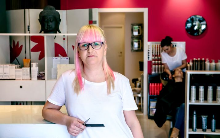 Hairdresser with pink hair in salon with client having hair washed in background