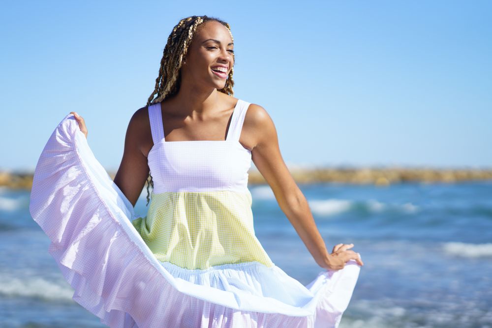 Black female holding up bottom of colorful dress while walking along the shore