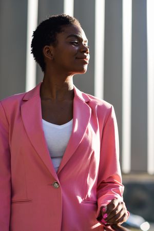 Female in suit with pink jacket outside office with striped shadows looking to her side, vertical