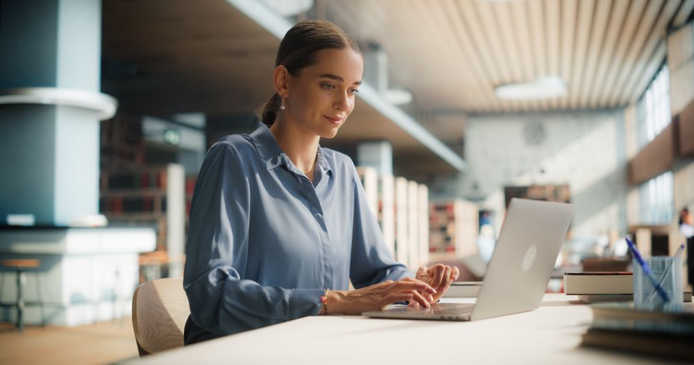 Female Student Engaged in Academic Research on Her Laptop in a Modern Library.
