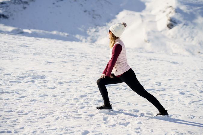 Side view of female in winter gear stretching legs on snowy mountain
