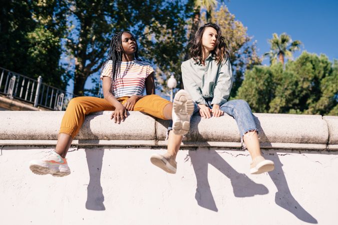 Serious female friends sitting on outdoor wall on sunny day