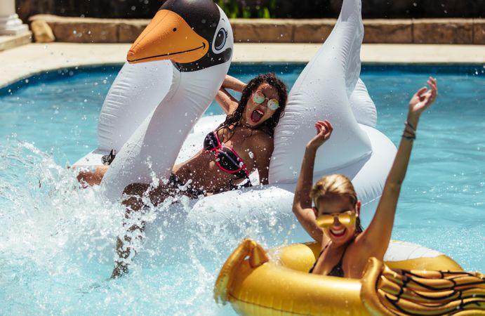 Two playful women splashing water while on pool floats
