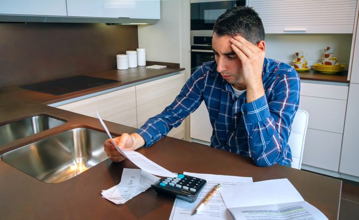 Stressed man using calculator while working with bills in kitchen