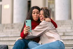 two students taking a selfie sticking out their tongues sitting on the stairs of the college.