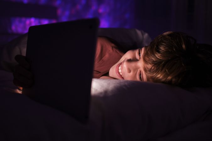 Cheerful kid browsing tablet in dark bedroom