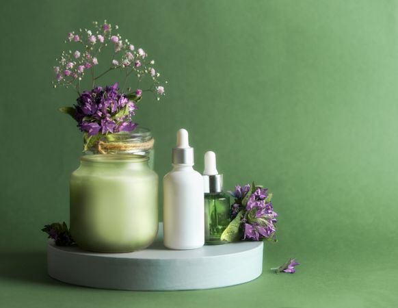 Natural cosmetic product setting with pipette bottle, candle and flowers on podium at green background.
