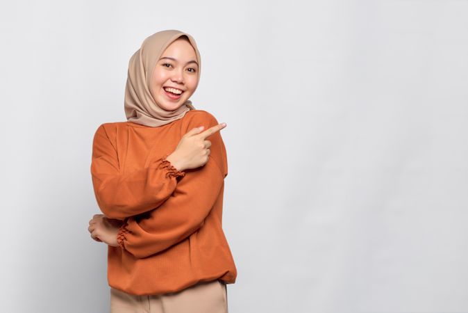 Smiling Muslim woman in headscarf and orange sweater pointing finger to blank copy space on her side