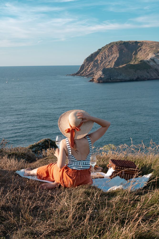 Woman with glass of drink on picnic mat looking at sea and mountains