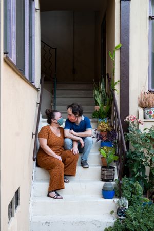 Romantic partners sitting on stairs for apartment looking at each other with masks on