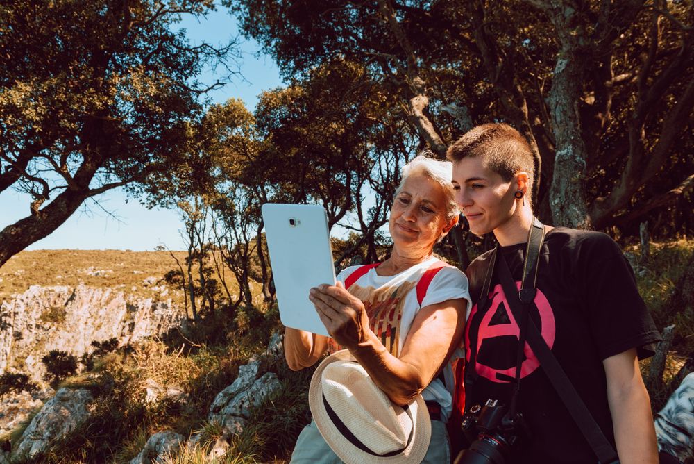Older woman and punk woman looking at tablet sitting on stone in valley