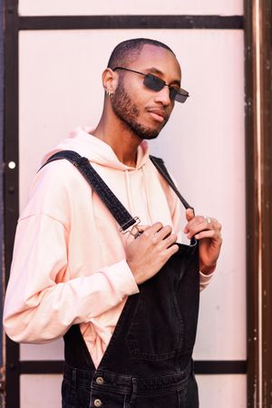 Serious male in denim overalls and earrings against a pink wall