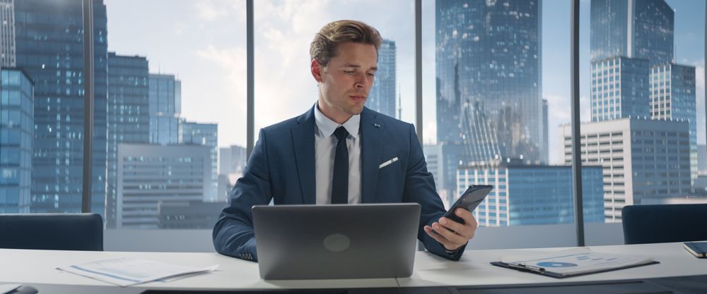 Confident Businessman in a Suit Sitting at a Desk in Office, Using Laptop Computer and Picking Up A Call