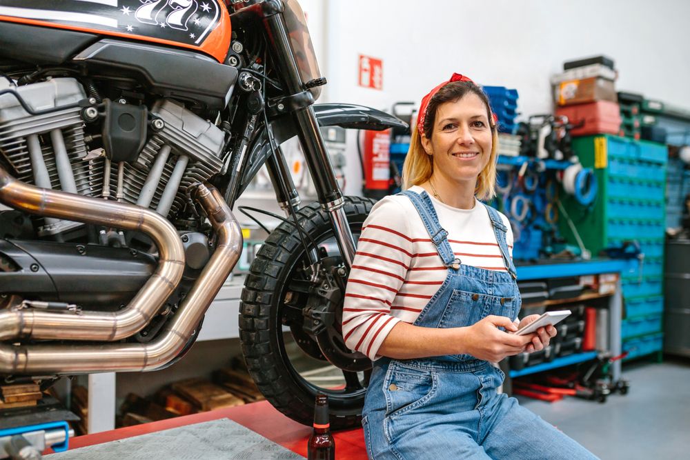 Mechanic woman holding phone sitting over platform with motorcycle on factory