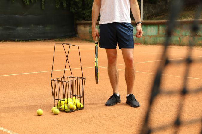 Man hold racket on clay court with basket of tennis balls