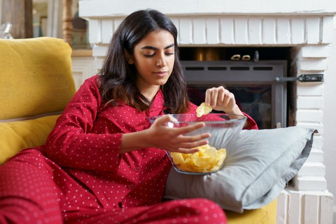 Female in red pajamas lying on sofa and snacking on chips