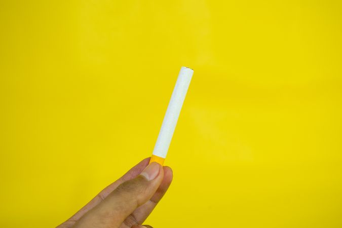 Fingers holding cigarette against yellow wall
