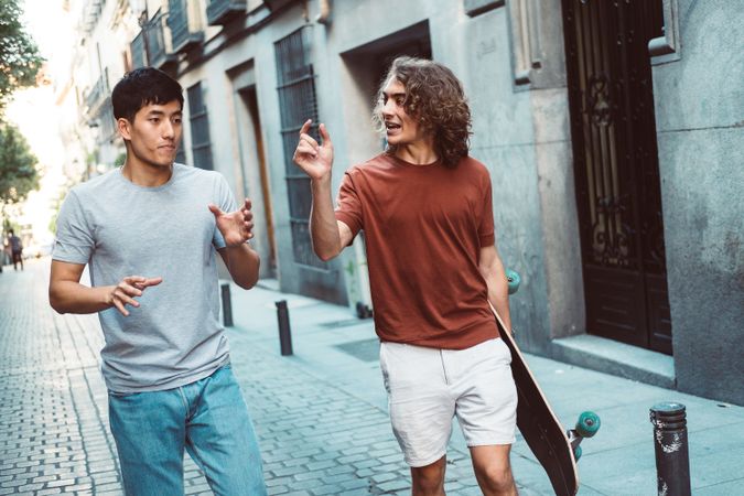 Cool multiracial male friends chatting while walking on street with longboard