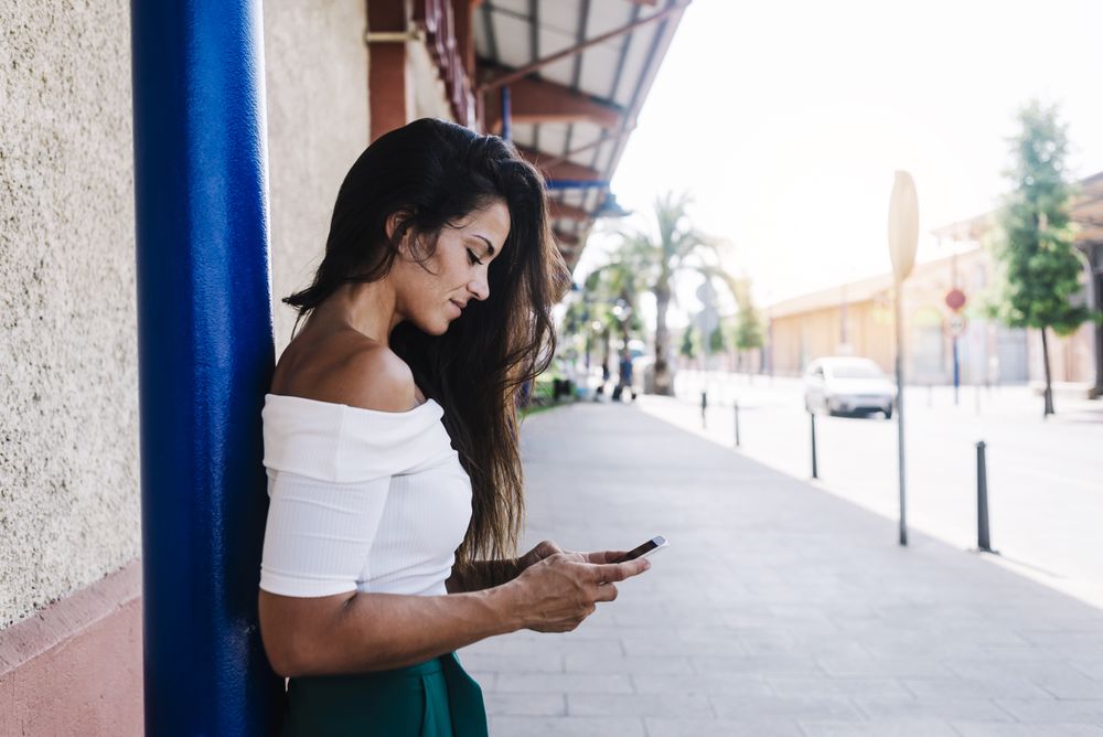 Happy young woman with long hair leaning against wall looking at cell phone