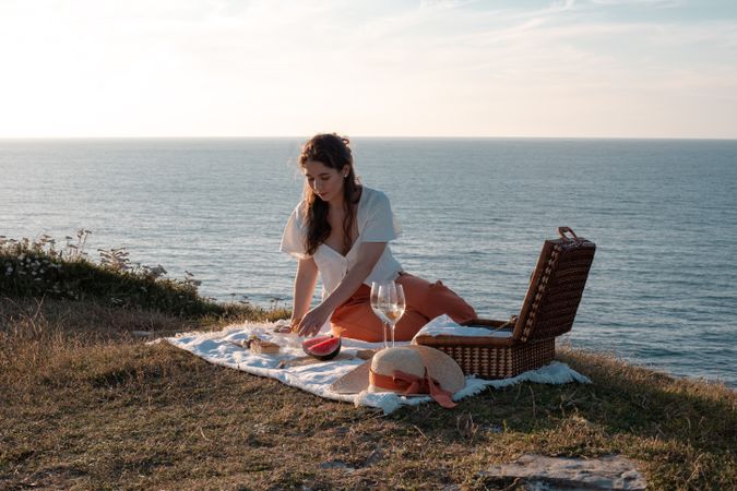 Woman with glass of drink on picnic mat near sea and mountains