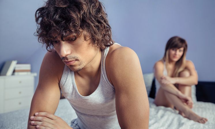 Upset man sitting on edge of bed after argument with woman in background