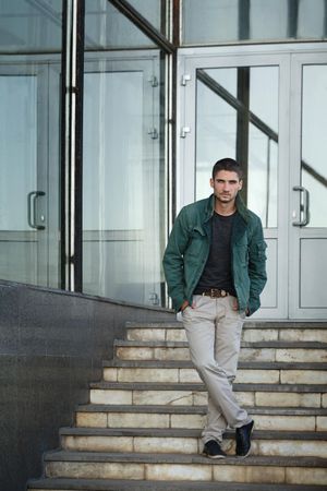 Male in green jacket standing with legs crossed on steps to building