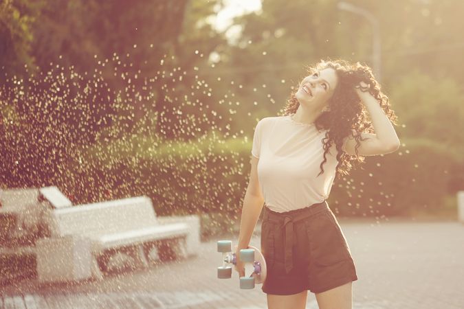 Happy female teenager with skateboard in park as sprinklers go off