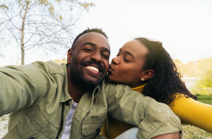 Black couple taking a selfie kissing on the cheek while sitting in a park