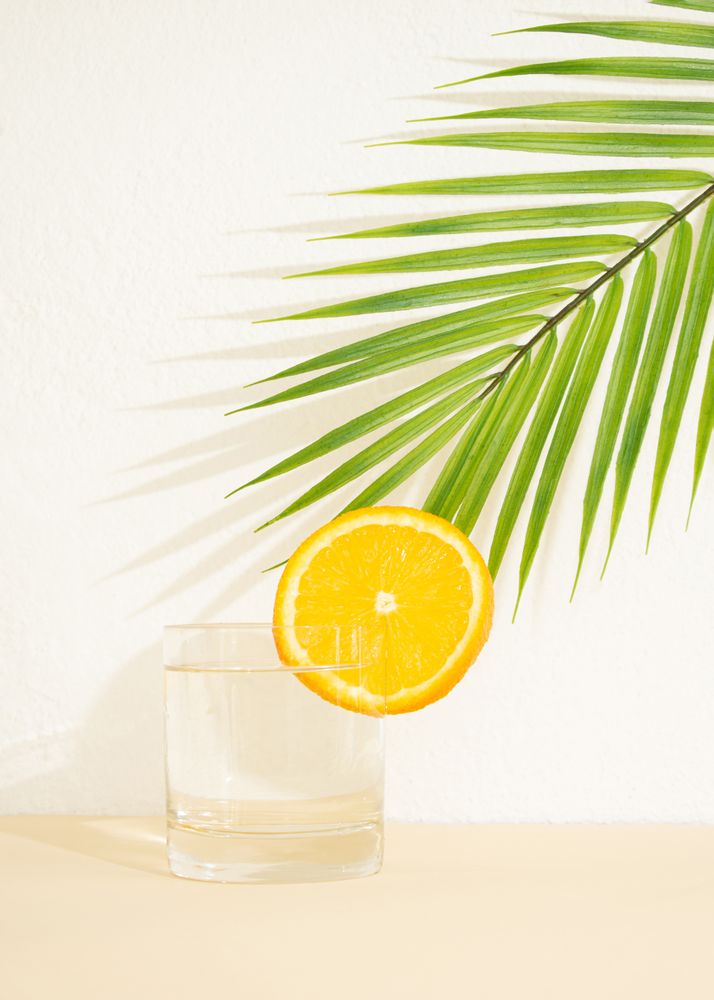 Glass of water with orange slide on neutral background and palm leaf