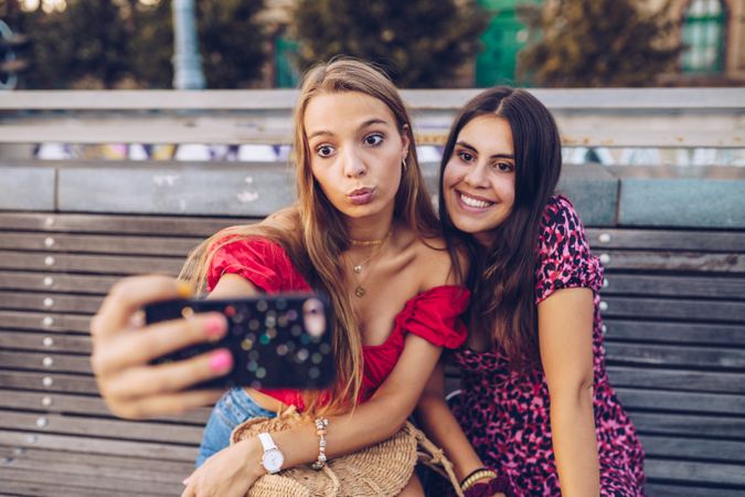 Laughing women taking selfie on wooden bench at street