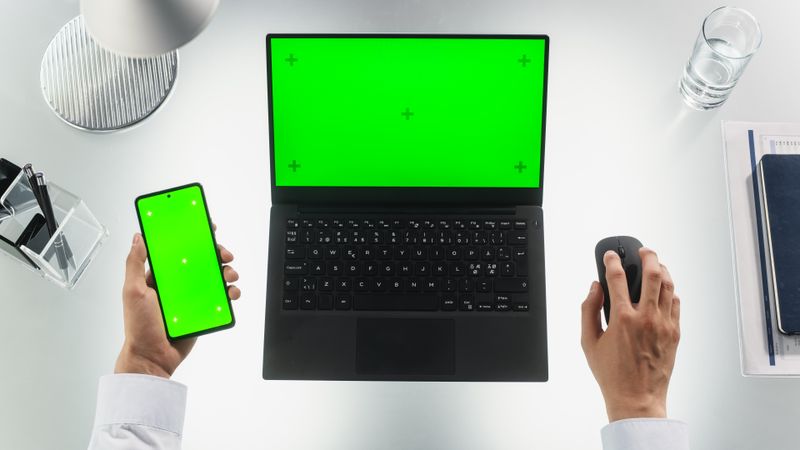 Top Down View of an Anonymous Person Multitasking with Laptop Computer and a Modern Smartphone. Both Devices are with Mock Up Green Screen Displays with Motion Tracker Templates. Man is Using a Mouse