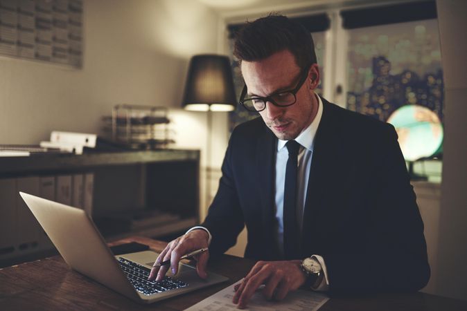 Man in suit and glasses working on his laptop and reading documents at night