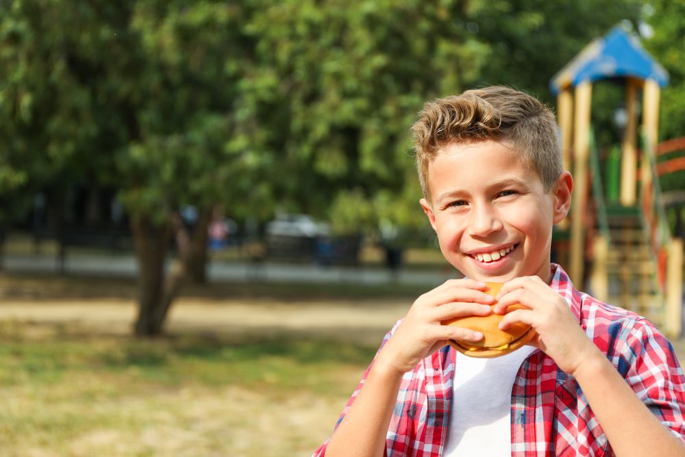 Boy with burger against blurred playground background, space for text