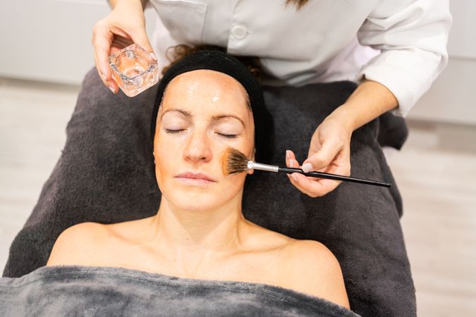 Beautician applying facial gel on face with brush during skin treatment in beauty spa