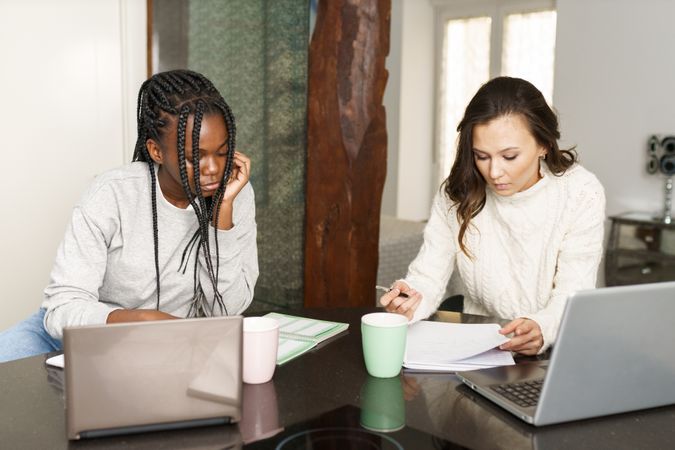 Two women sitting at kitchen table doing work on laptops