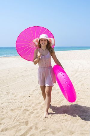 Beautiful woman in pink dress walking on sand beach wearing a hat and holding a parasol and a floater