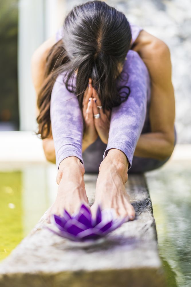 Aesthetic shot of a praying woman sitting behind a purple glass lotus on a tree trunk over the water.