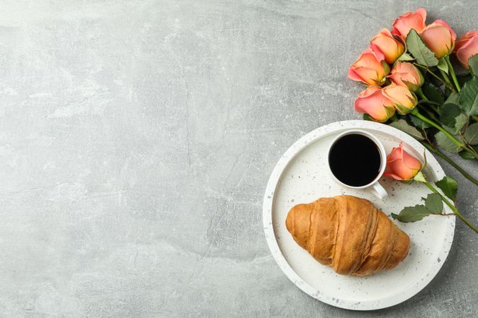 Composition with marble tray, cup of coffee, croissant, roses and space for text on grey background, top view