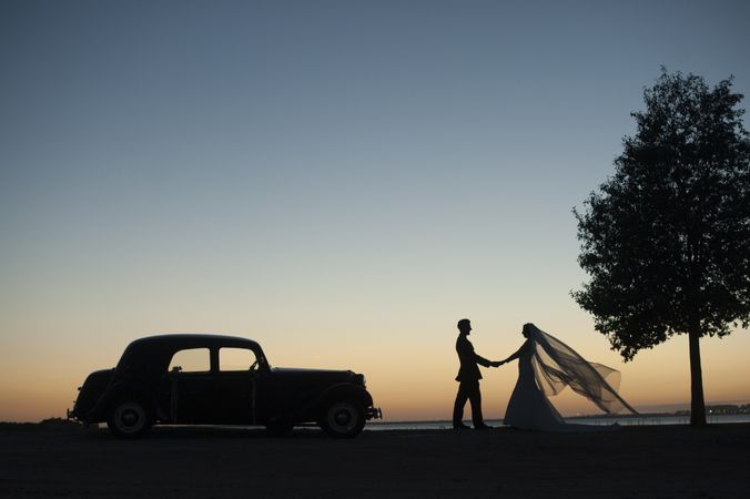 Woman and man holding hands near vintage car at sunset