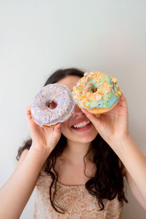 Playful woman making glasses of donuts while standing by white wall