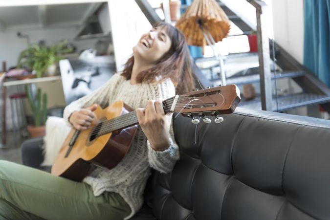 Happy female on sofa playing acoustic guitar and singing at home in living room