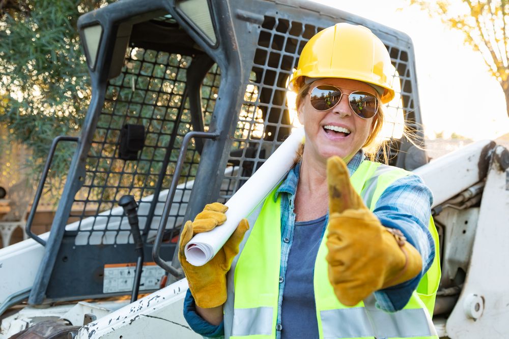 Smiling Female Worker With Thumbs Up and Technical Blueprints Near Small Bulldozer At Construction Site