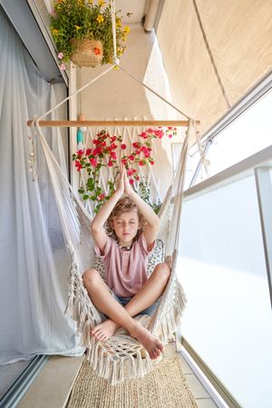 Concentrated boy sitting in hammock chair and making namaste gesture