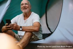 Smiling mature man sitting in tent with his mobile phone 4MKDx0
