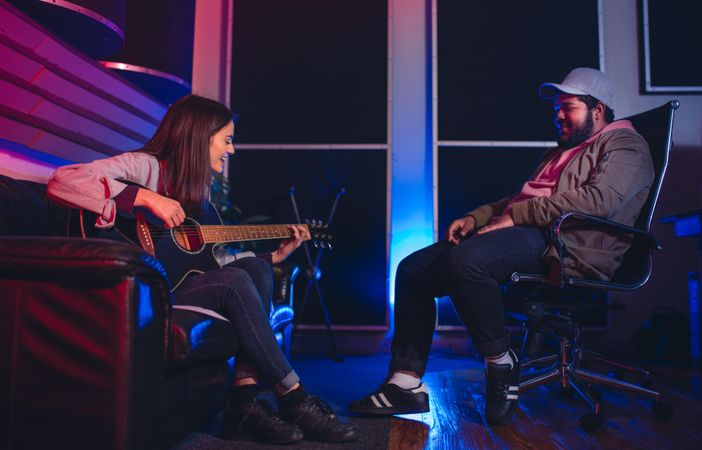 Woman playing guitar and singing with man sitting on chair listening in recording studio