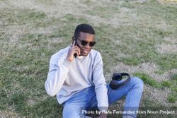 Happy Black male with sunglasses sitting in the grass talking on his phone 0gXaRN